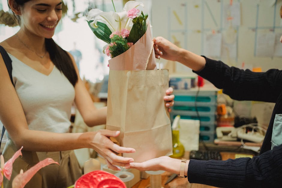A woman in a white tank top is smiling as she receives a bouquet of fresh flowers from a person in dark clothing inside a flower shop. The bouquet, wrapped in light pink paper, contains white calla li