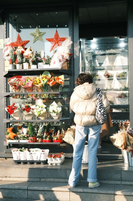 A woman with curly dark hair, dressed in a beige puffer jacket and light blue jeans, stands outside a flower and gift shop on a sunny day. She faces the shop window, which displays various floral arra