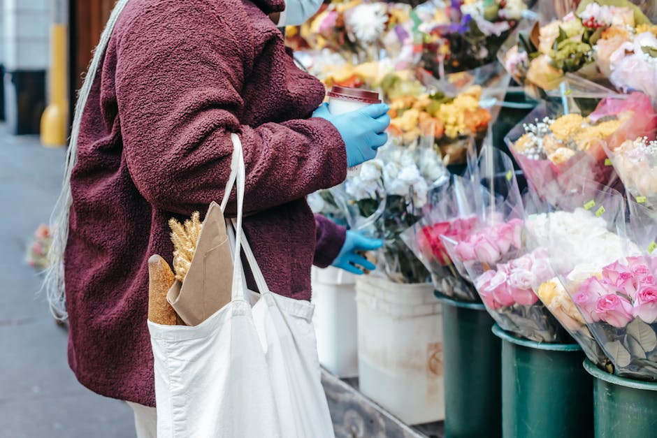 A person wearing a burgundy fleece jacket and blue gloves is shopping for flowers at an outdoor floral market. They are holding a white shopping tote with a brown paper bag and a baguette inside, hang