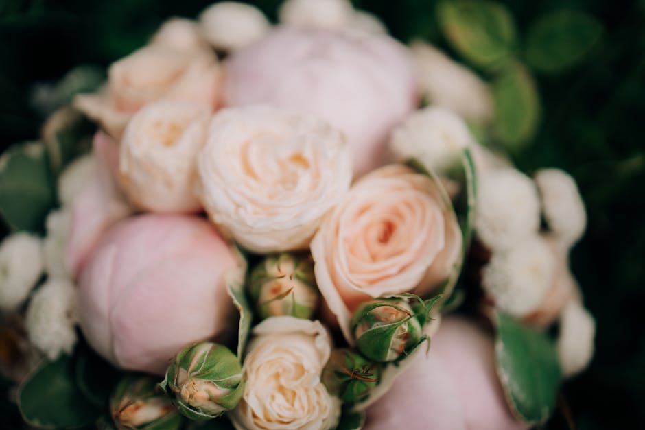 A close-up image of a floral arrangement featuring pale peach and soft pink roses with tightly packed, layered petals, complemented by small white filler flowers and green leaves. The bouquet has a na
