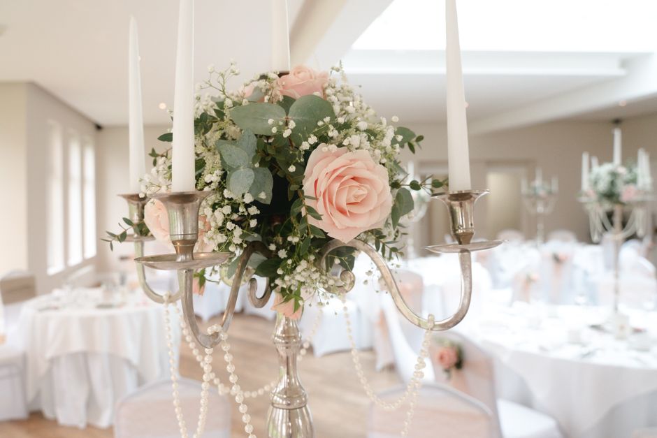 A floral arrangement featuring soft pink roses, white baby's breath, and eucalyptus leaves, elegantly displayed on a silver candelabrum with three candle holders. The bouquet is lush and fresh, with d