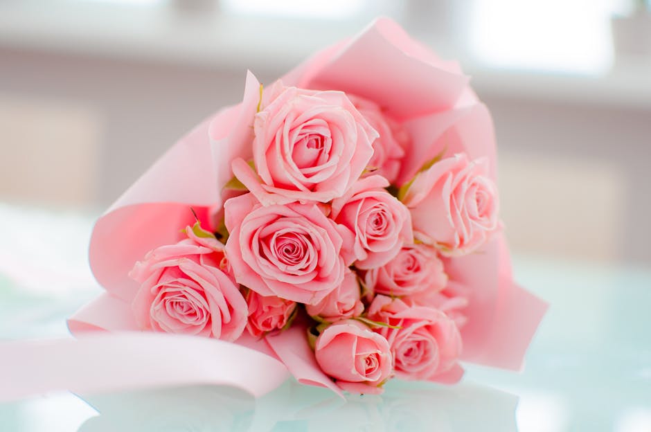 A bouquet of fresh pink roses arranged with their stems wrapped in light pink paper, forming a rounded floral display placed on a smooth, reflective surface. The roses, with tightly furled petals, app