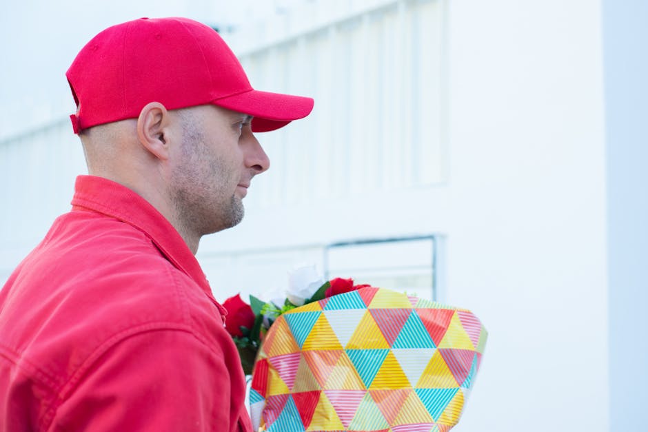 A man dressed in a red cap and matching red jacket is holding a wrapped floral gift, featuring a vibrant geometric pattern with triangles in shades of yellow, blue, red, and pink. The flowers are nest
